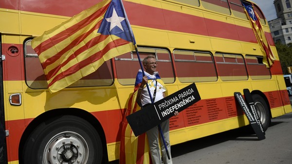 A man poses with a sign reading "Next stop: Independence" and an "Estelada" (pro independence Catalan flag) past a bus with the colours of the Catalan flag